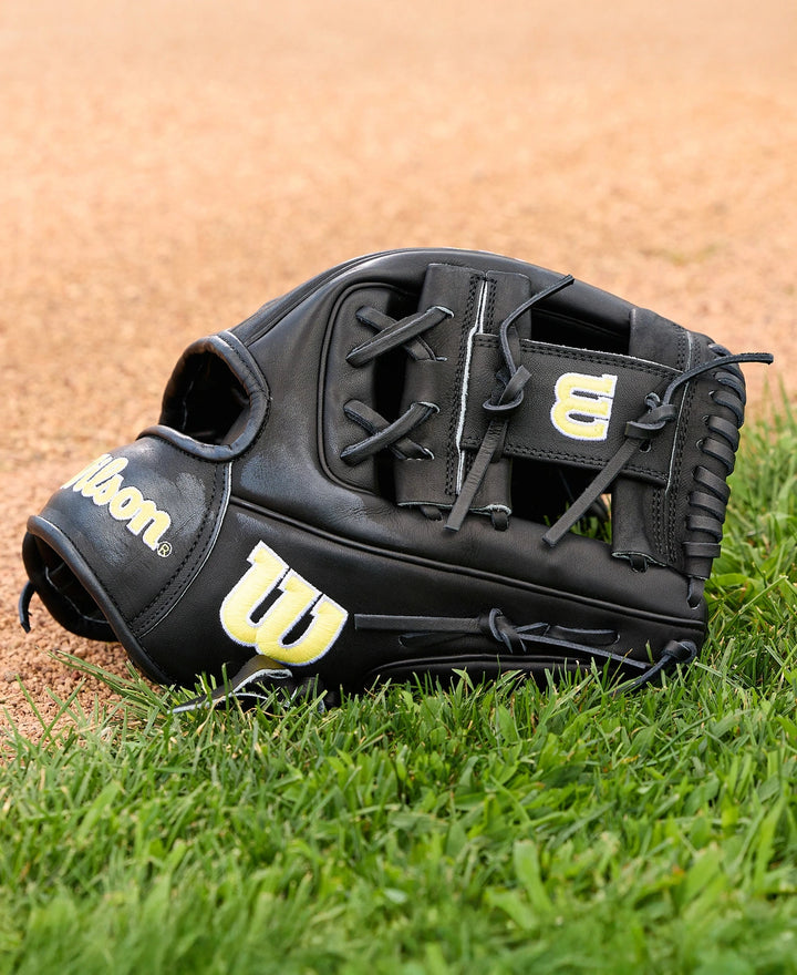 A Wilson A2000 1788 11.25" baseball glove by Wilson, featuring a black design with yellow and white logos, rests on green grass near the dirt infield. Crafted from premium Pro Stock Leather.