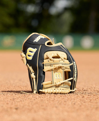 A Wilson A2000 1786 11.5" Baseball Glove (WBW104108115), featuring H-Web design in black and tan, sits upright on a sandy baseball field, with blurred green outfield fencing and trees in the background.