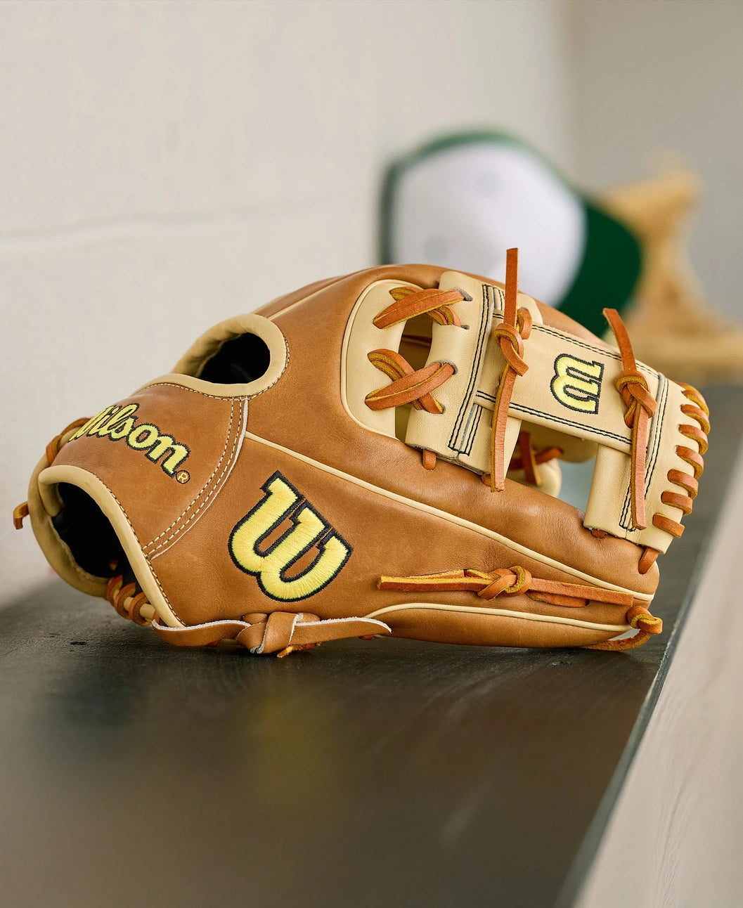 A Wilson A2000 1786 11.5" Baseball Glove (WBW104109115) with tan and cream Pro Stock Leather and brown laces sits on a dark shelf, while a blurred green and white cap appears in the background.