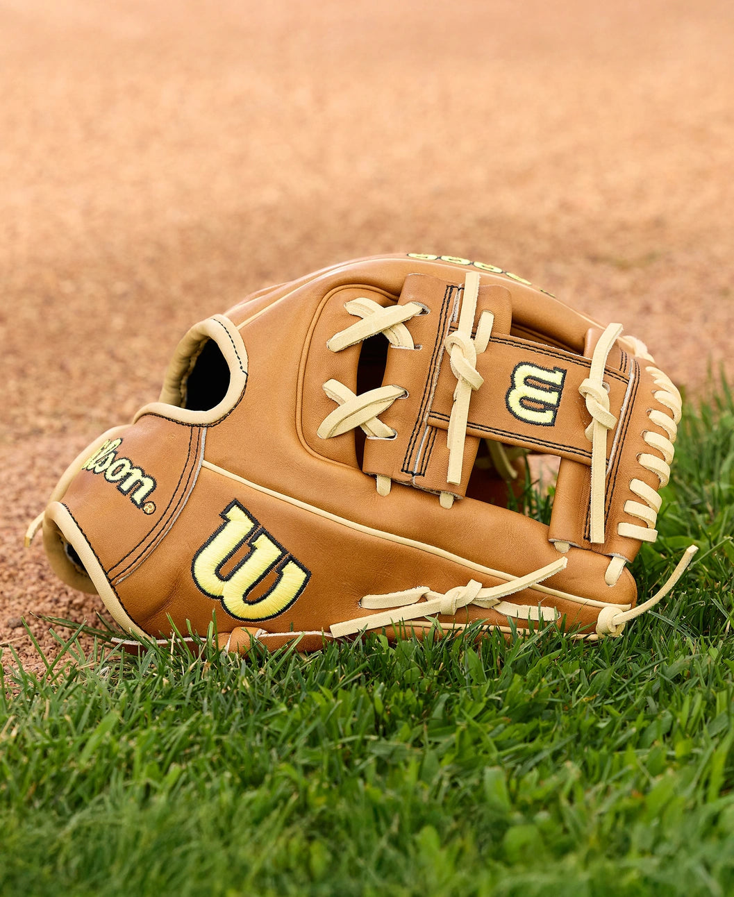 A Wilson A2000 DP15 11.5" Baseball Glove by Wilson, with tan Pro Stock Leather and cream laces, rests on green grass beside a dirt infield, ready for action.