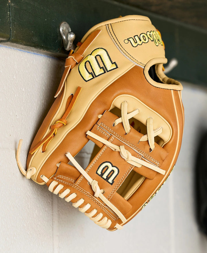 A tan and light brown Wilson A2000 1975 11.75" Baseball Glove (WBW1041131175) hangs on a metal hook attached to a white wall, with the Wilson logo visible on the thumb and wrist straps.