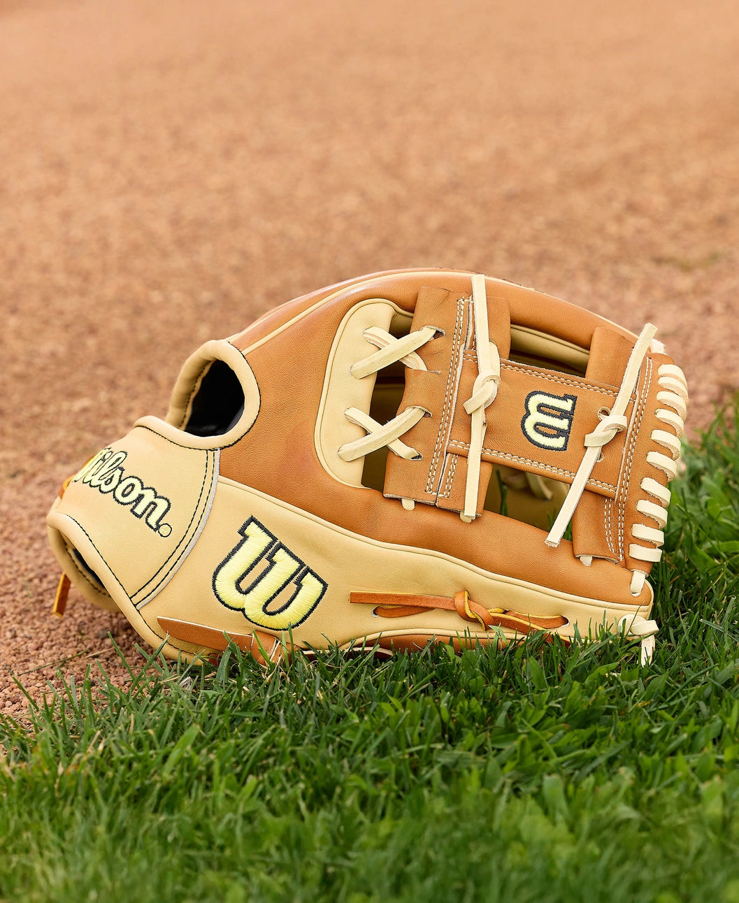 A Wilson A2000 1975 11.75" Baseball Glove (WBW1041131175) in tan and cream, made with Pro Stock Leather, sits on green grass near a dirt infield. The Wilson logo stands out on this infield glove.