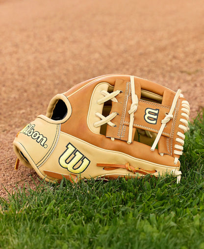 A Wilson A2000 1975 11.75" Baseball Glove (WBW1041131175) in tan and cream, made with Pro Stock Leather, sits on green grass near a dirt infield. The Wilson logo stands out on this infield glove.