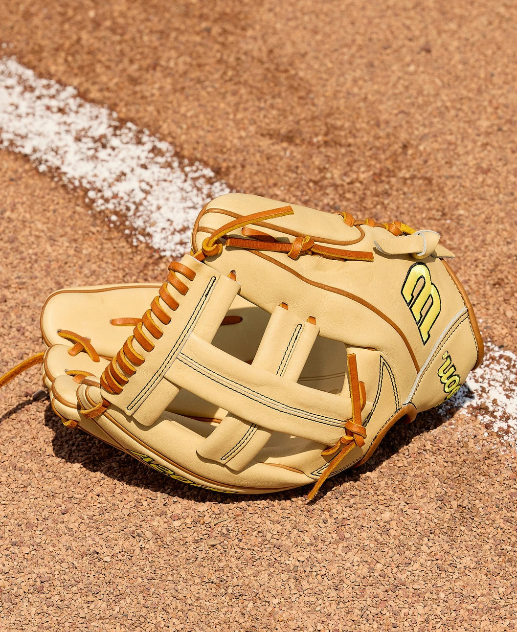 A Wilson A2000 EL3 11.75" Baseball Glove (WBW1041141175) by Wilson, featuring a tan design with brown laces, lies open on sandy dirt beside a white chalk foul line on the baseball field.