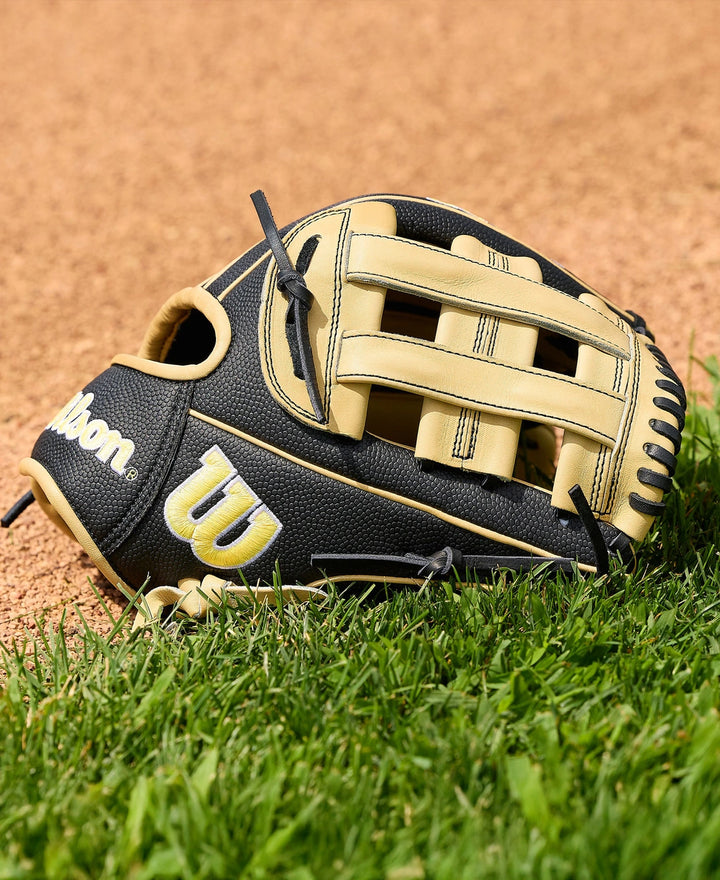 A Wilson A2000 DW5 12" baseball glove by Wilson, crafted from Pro Stock Leather in black and tan, lies on green grass near the edge of a dirt baseball field.