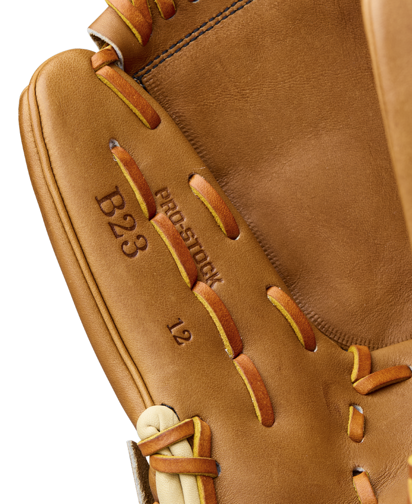 Close-up of a Wilson A2000 B23 12" Baseball Glove with brown Pro Stock leather, tan and orange laces, visible stitching, and embossed text: B23, PRO-STOCK, and 12. Model: WBW10411712.