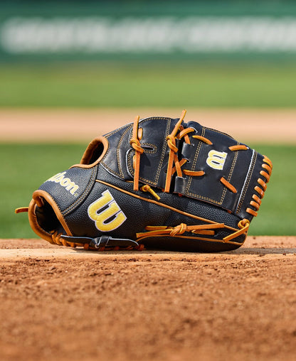 A Wilson A2000 B125SS 12.5" baseball glove (WBW104119125) by Wilson, in black and tan, sits on the dirt of a baseball field with the green outfield blurred in the background.