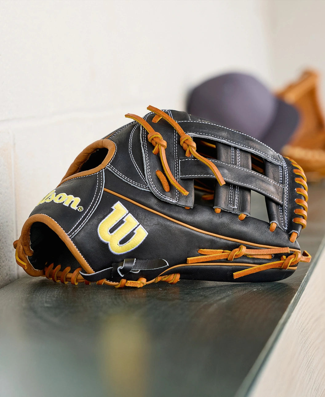 A Wilson A2000 1750 12.5" Baseball Glove (WBW104121125) by Wilson, featuring black and brown leather with yellow stitching and a "W" logo, rests on a metal bench next to a blurred hat and another glove in the background.