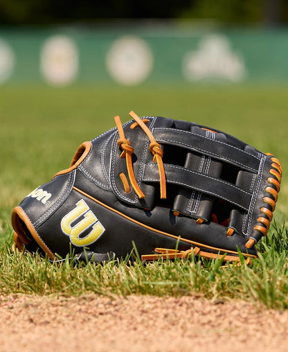 A Wilson A2000 1750 12.5" Baseball Glove (WBW104121125) by Wilson in black and brown leather lies on green grass near the dirt of a baseball field, with a blurred green fence and white signage in the background.
