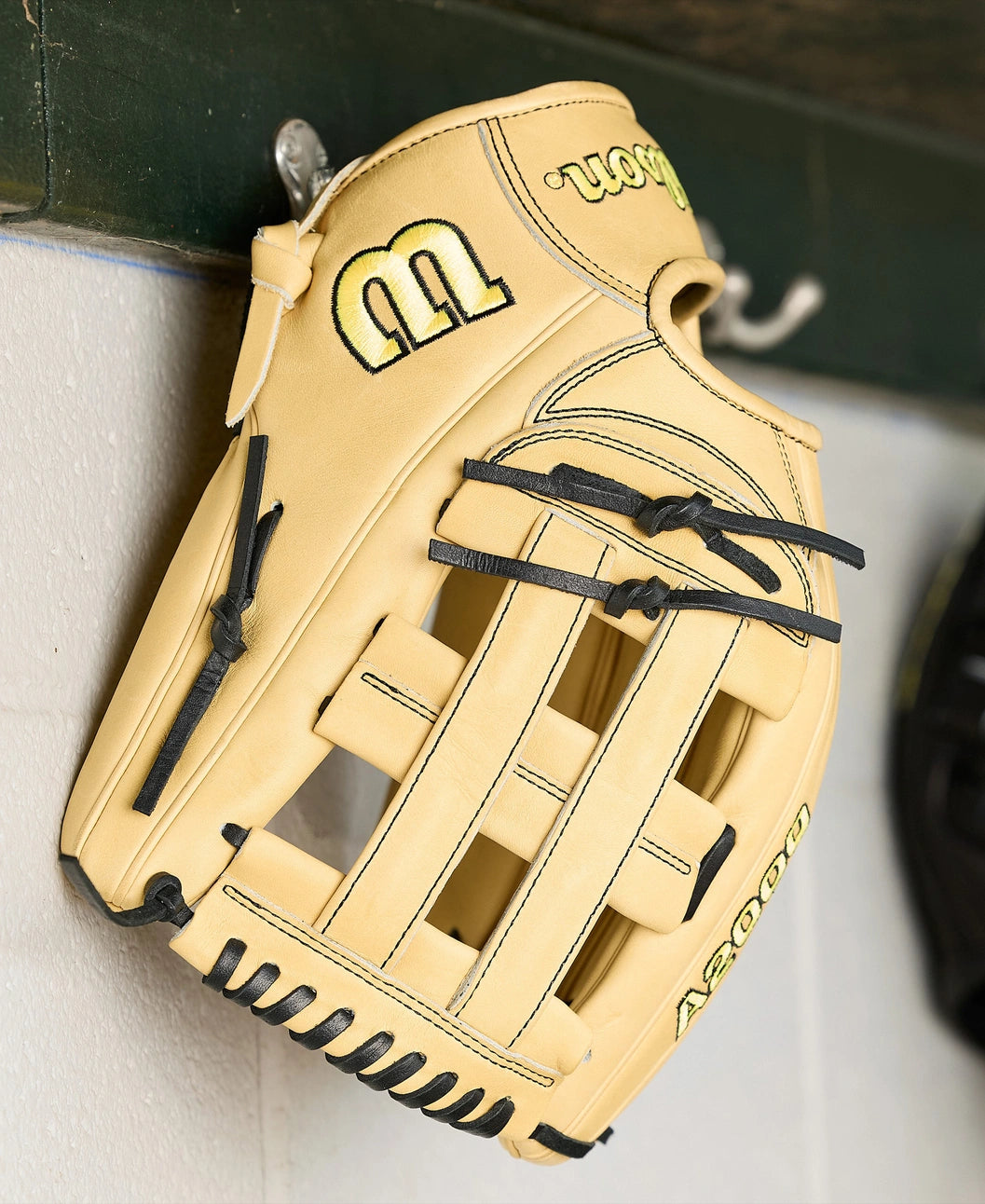 A Wilson A2000 1777 12.75" Baseball Glove (WBW1041231275) in tan with black laces, made from Pro Stock Leather, hangs on a metal hook against a white wall with a green shelf above.