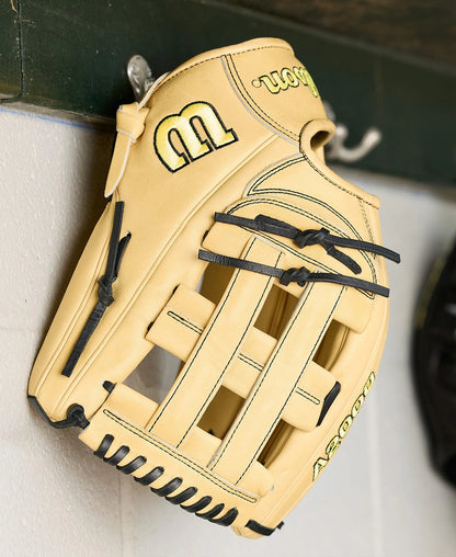 A Wilson A2000 1777 12.75" Baseball Glove (WBW1041231275) in tan with black laces, made from Pro Stock Leather, hangs on a metal hook against a white wall with a green shelf above.