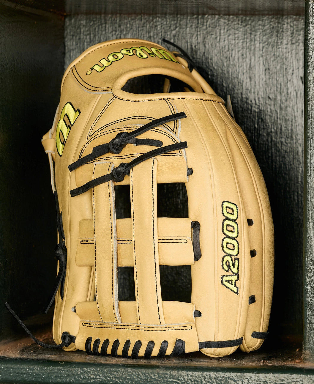 A Wilson A2000 1777 12.75" Baseball Glove (WBW1041231275) in tan with black laces and yellow lettering, made from Pro Stock Leather, stands upright in a dark green wooden cubby.