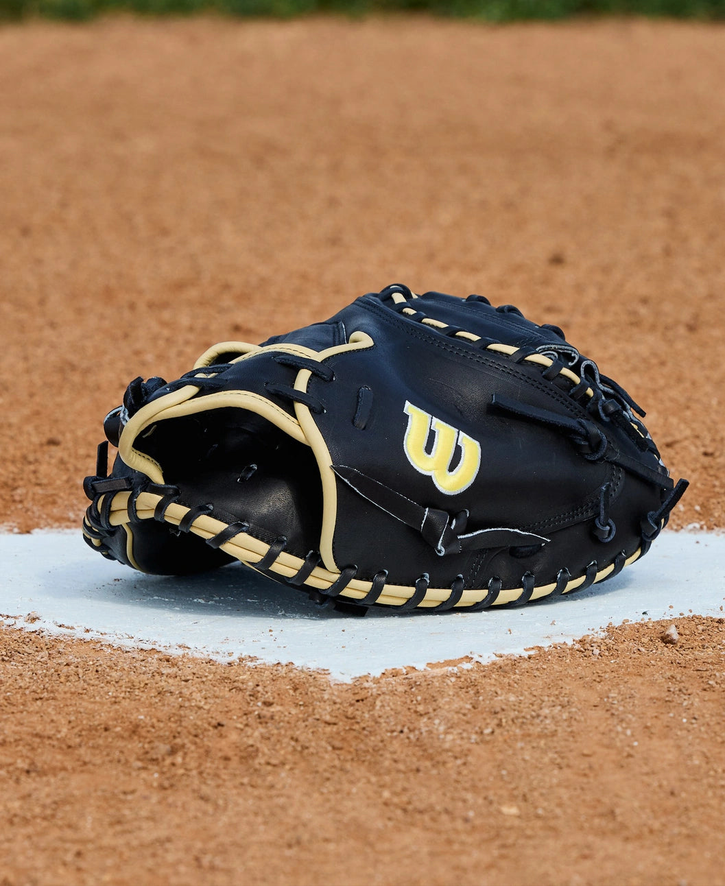 A Wilson A2000 1734 34" Baseball Catcher’s Mitt (WBW10412834), black and tan with a yellow W logo, rests on home plate of a dirt baseball field with grass in the background.