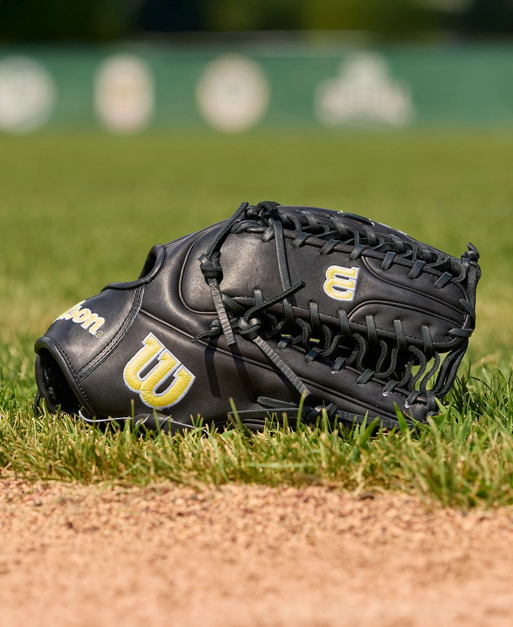 A Wilson A2000 OT7 12.75" Baseball Glove (WBW1041811275) by Wilson, crafted with Pro Stock Leather, sits on green grass near a dirt infield, set against a blurred baseball field background.