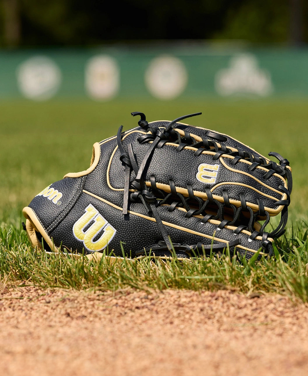A Wilson A2000 OT7SS 12.75" Baseball Glove (WBW1041831275) by Wilson rests on the grass near the infield dirt, with a blurred green outfield fence and logos in the background.