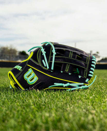 A Wilson A2000 1750SS 12.5" baseball glove (WBW104599125) in black with neon green and blue accents rests on grass, with a blurred baseball field and sky in the background.