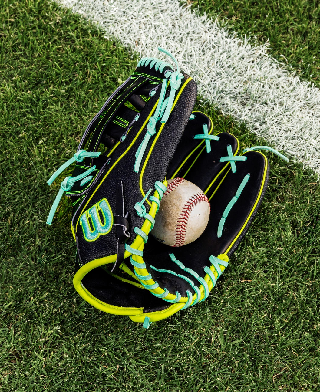 A Wilson A2000 1750SS 12.5" Baseball Glove (WBW104599125) in black with neon green and blue laces rests open on grass near a white line, holding a baseball. Made with premium Pro Stock leather for lasting performance.