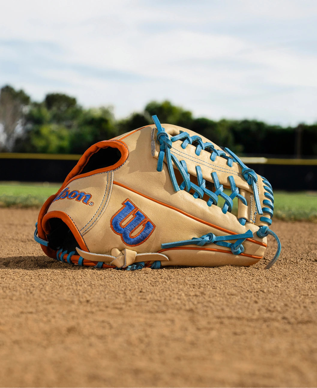 A Wilson A2000 PF89 11.5" baseball glove with tan Pro Stock leather and blue and orange laces lies on a dirt infield, green grass and trees in the background.