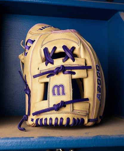 A Wilson A2000 H12 12" Fastpitch Glove (WBW10460712) in tan with purple laces and stitching, crafted from Pro Stock Leather, displays "m" and "A2000" lettering as it rests on a blue shelf.