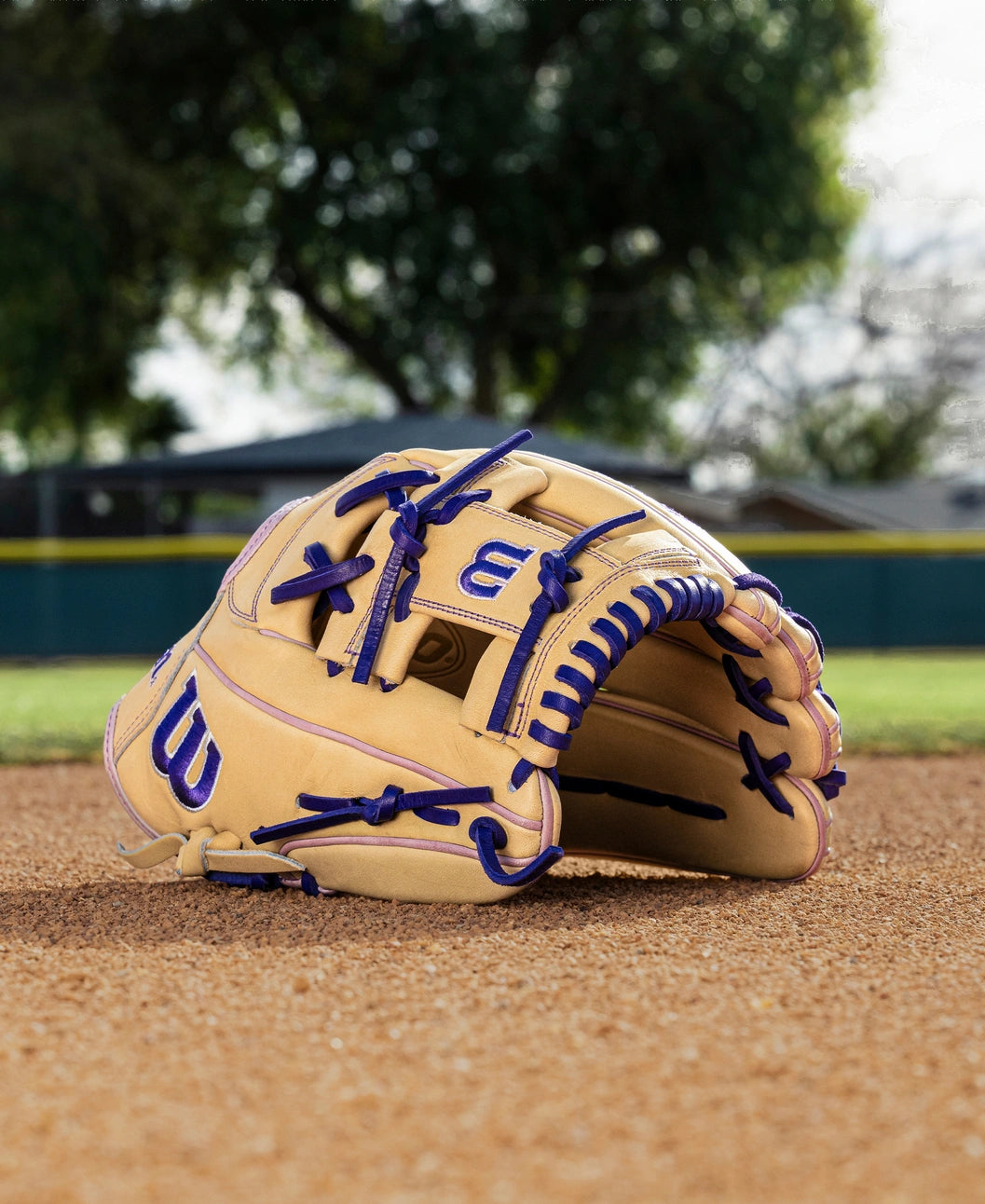 A Wilson A2000 H12 12" Fastpitch Glove (WBW10460712) in tan with blue laces lies on a dirt infield, with trees, a fence, and a house softly blurred in the background.