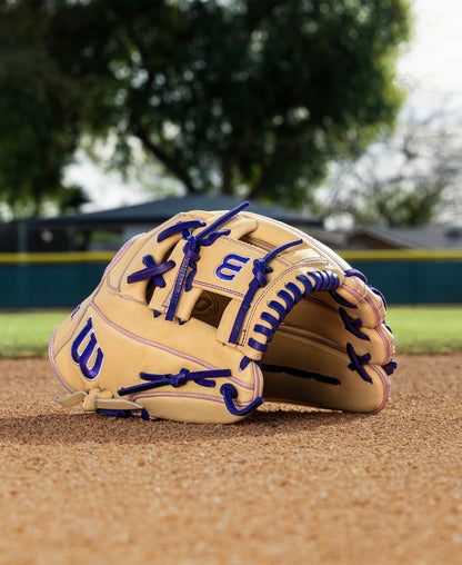 A Wilson A2000 H12 12" Fastpitch Glove (WBW10460712) in tan with blue laces lies on a dirt infield, with trees, a fence, and a house softly blurred in the background.