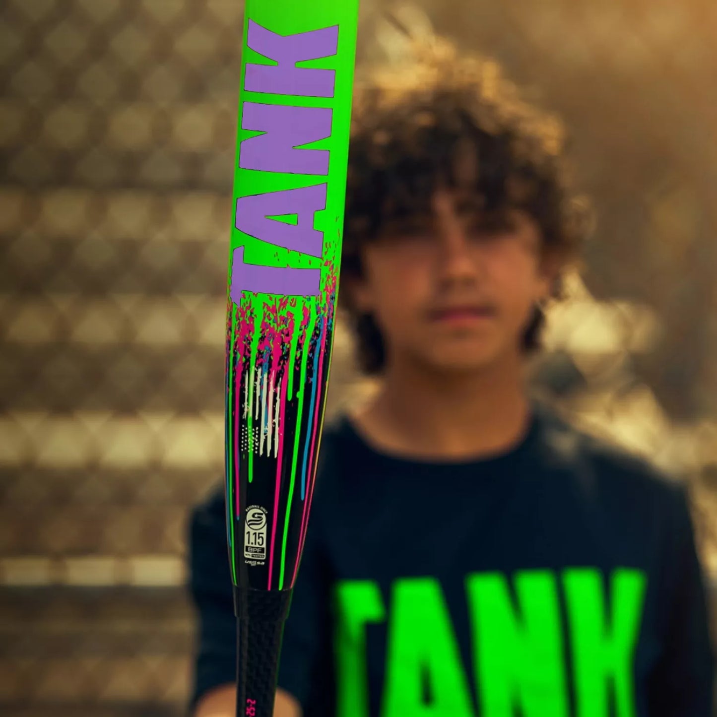 A boy, blurred in the background, holds a sharply focused Soldier Sports 2026 Soldier Tank Double Wall (-8) 2 3/4" USSSA Baseball Bat (Y-8-26-2), featuring neon green and purple accents. He wears a black “TANK” shirt behind a chain-link fence.