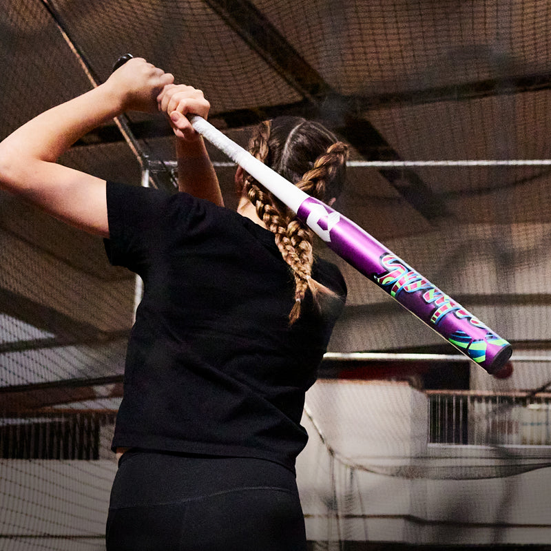 A person with braided hair, in a black outfit, swings the 2025 DeMarini Spryte (-12) Fastpitch Softball Bat (WBD2553010) by DeMarini inside an indoor batting cage.