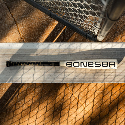 A white and black Warstic 2026 Bonesaber Hybrid (-10) USA Baseball Bat rests on a metal bench near a chain-link fence, with sunlight casting shadows on the ground and bench.