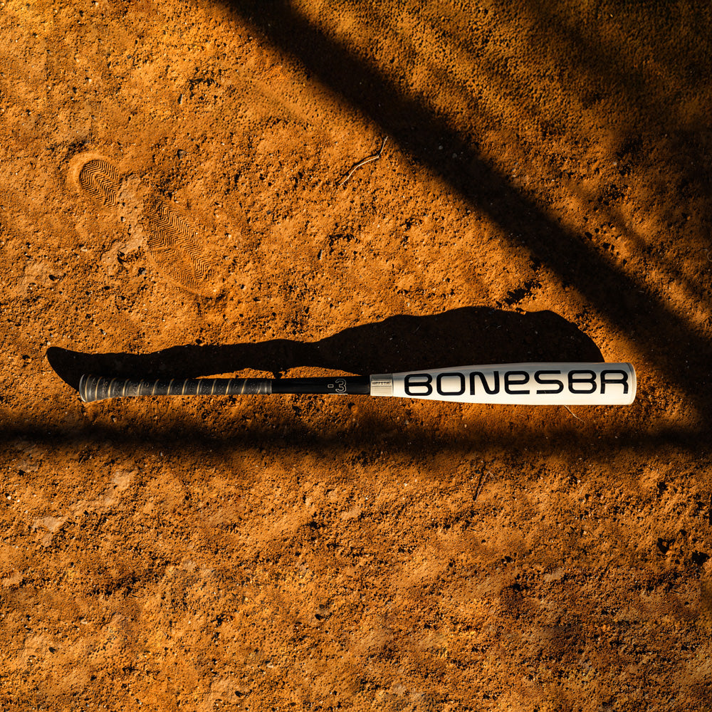 A 2025 Warstic Bonesaber Hybrid (-3) BBCOR Baseball Bat (MBBSRHB25WH3) lies on reddish-brown dirt, casting a long shadow in the sunlight as dark diagonal shadows cross the ground.