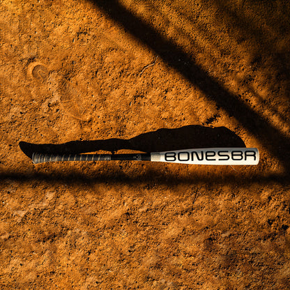 A 2025 Warstic Bonesaber Hybrid (-3) BBCOR Baseball Bat (MBBSRHB25WH3) lies on reddish-brown dirt, casting a long shadow in the sunlight as dark diagonal shadows cross the ground.
