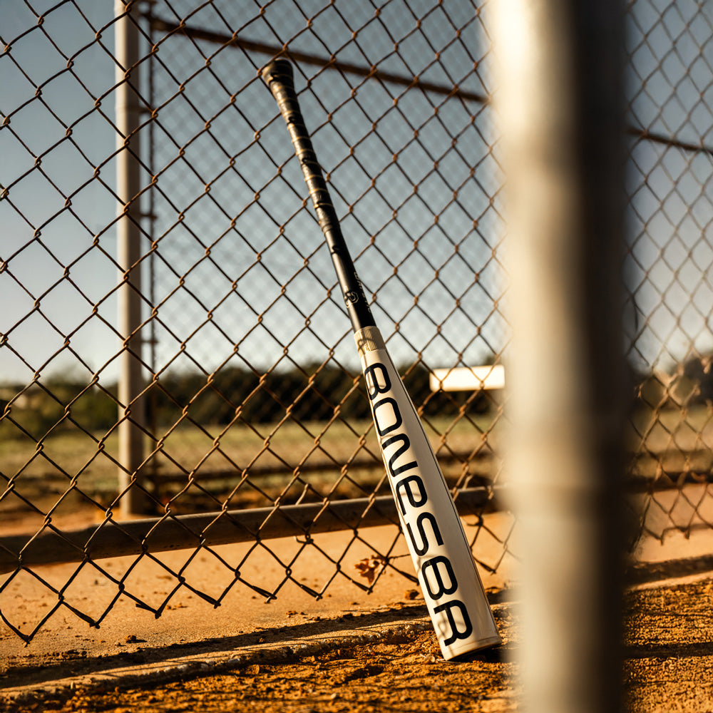 A 2025 Warstic Bonesaber Hybrid (-3) BBCOR Baseball Bat (MBBSRHB25WH3) rests against a chain-link fence on a sandy field, sunlight casting long shadows—highlighting the premium quality of a top-tier Warstic BBCOR bat.