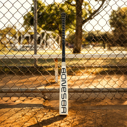The 2025 Warstic Bonesaber Hybrid (-3) BBCOR Baseball Bat (MBBSRHB25WH3) stands upright in black and white against a chain-link fence, with trees and a sunlit park in the background.