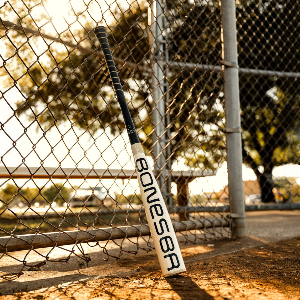 A 2025 Warstic Bonesaber Hybrid (-3) BBCOR Baseball Bat (MBBSRHB25WH3) leans against a chain-link fence by a sunlit baseball field, highlighting Warstic’s signature style amid benches and trees in the background.