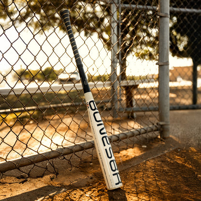 A white and black Warstic 2026 Bonesaber Hybrid (-10) USA Baseball Bat leans against a chain-link fence on a dirt field with trees and sunlight in the background.
