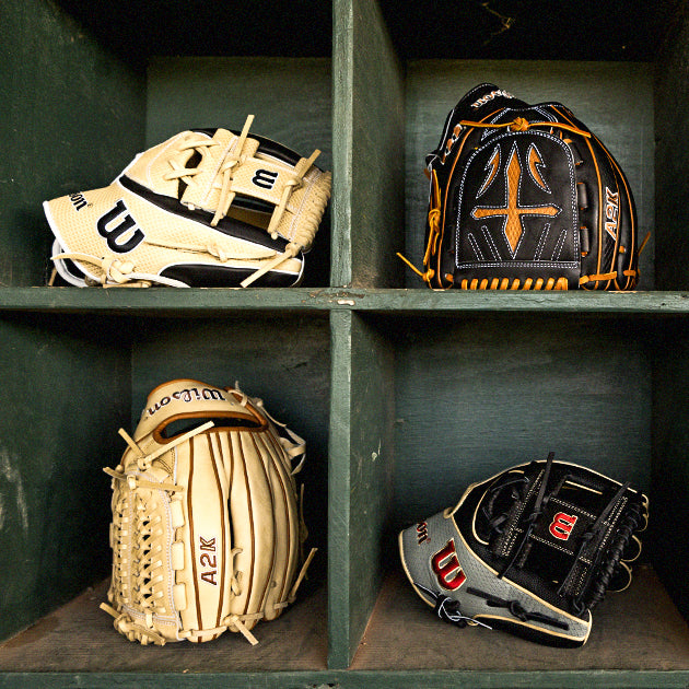 Four baseball gloves, two tan and two black, are neatly placed in separate cubby holes of a wooden shelf, showing different brands and stitching styles.