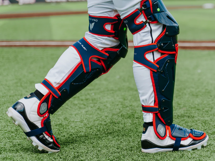 Close-up of a baseball catchers legs wearing white pants, blue and red protective shin guards, and cleats, standing on green artificial turf.