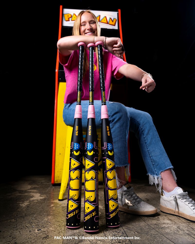 A smiling person in a pink shirt and jeans sits in front of a yellow PAC-MAN arcade machine, leaning on three Marucci 2027 ASURA Rise PAC-MAN (-10) Fastpitch Softball Bats featuring Rise Sync Barrel Technology.