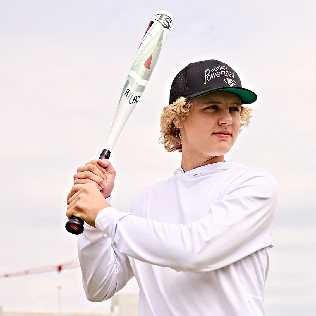 A young person with curly blond hair in a black hat and white long-sleeve shirt holds the 2025 Louisville Slugger Atlas (-5) 2 5/8" USSSA Baseball Bat (WBL2981010), preparing to swing outdoors under a cloudy sky.