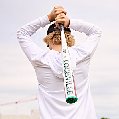 A blonde person in a white hoodie and black cap stands outdoors, facing away while holding a Louisville Slugger 2025 Atlas (-5) 2 5/8" USSSA Baseball Bat (WBL2981010) over their shoulders.