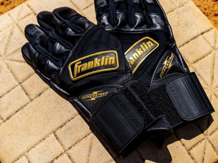 A pair of black Franklin brand batting gloves with gold lettering and detailing rest on a tan baseball base.