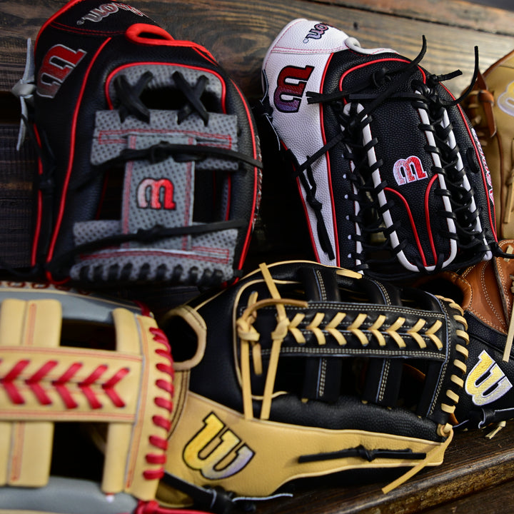 A close-up of several colorful baseball gloves with different stitching and patterns, all branded with the Wilson W logo, arranged on a wooden surface.