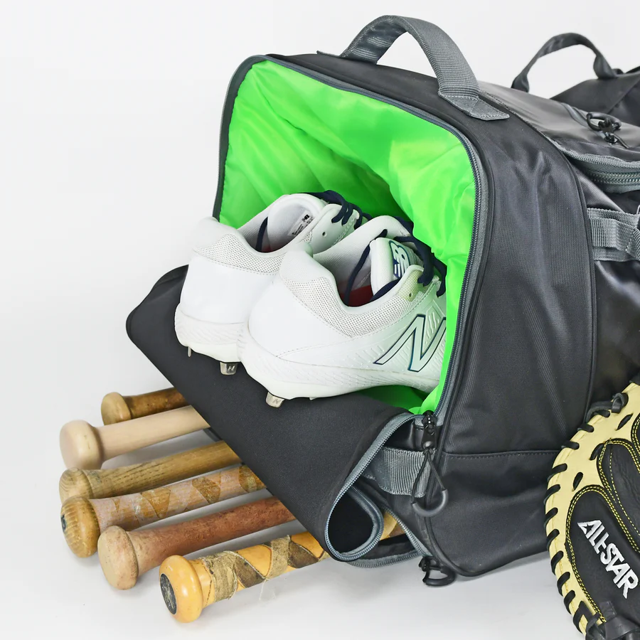 The All Star S7 Elite Catcher's Roller Bag (BB-S7E-RB) with a bright green interior holds white athletic shoes. Several wooden baseball bats and a black and tan glove are partially visible next to the bag.