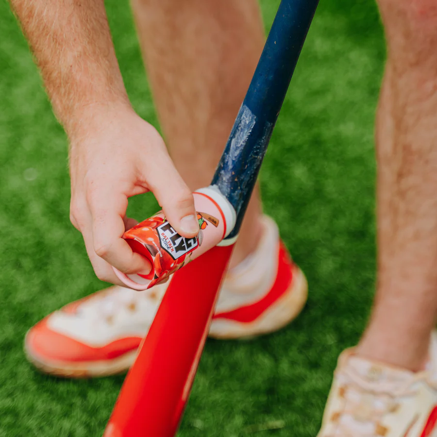 A person applies the All Star Big Fly Bat Tack Stick (BFST1) to a baseball bat for extra grip while standing on green grass, wearing red and white athletic shoes.