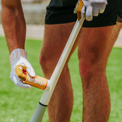 A person in gloves and shorts applies the All Star Big Fly Bat Tack Stick (BFST1) to a garden tool handle while standing on grass.