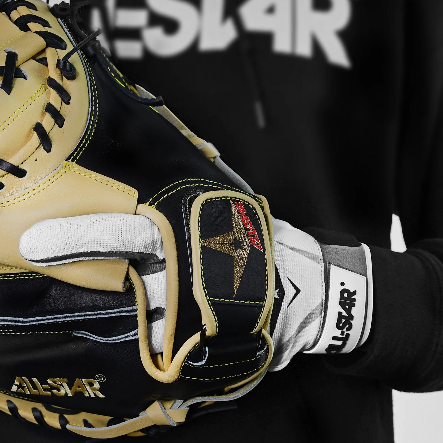 A close-up of a person wearing the All Star S7 Axis Padded Inner Glove (CG6001) and holding a black and tan baseball glove, with part of an All Star logo visible on a black hoodie in the background.
