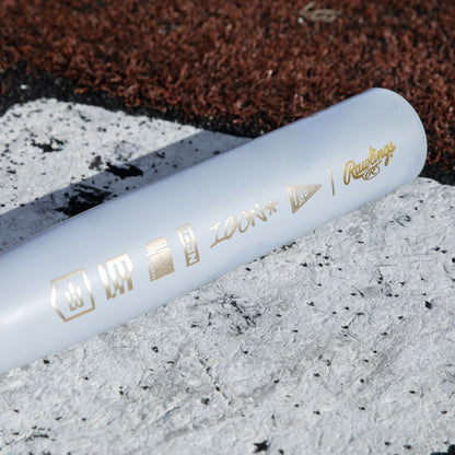 A close-up of the 2026 Rawlings Icon The Chosen One (-3) BBCOR Baseball Bat, featuring gold icons and "ICON" text, lies on a white and brown turf field.