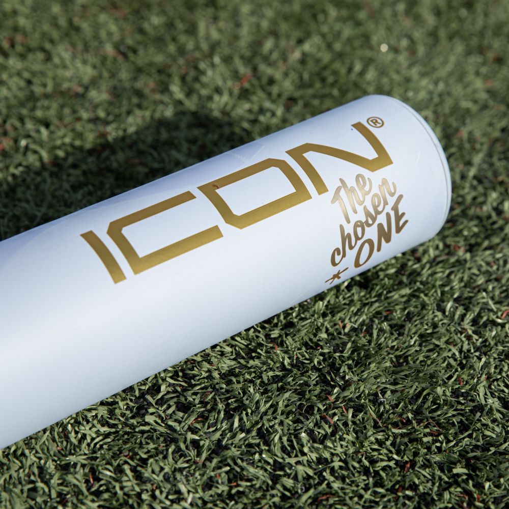 A white Rawlings 2026 Icon The Chosen One (-3) BBCOR Baseball Bat (RBB6IONE3) with gold "ICON The chosen ONE" lettering rests on artificial grass.