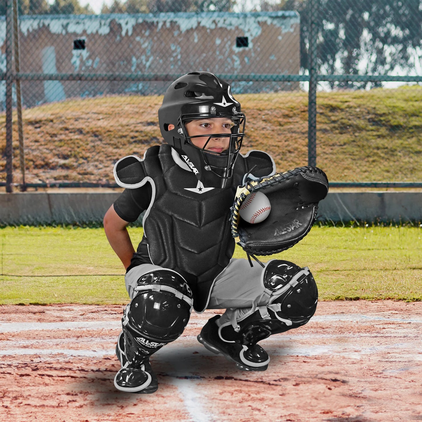 A young catcher wearing the All Star Future Star Series Catcher's Kit (CK-FS2) crouches behind home plate, chest protector on and glove ready, poised for the next pitch.