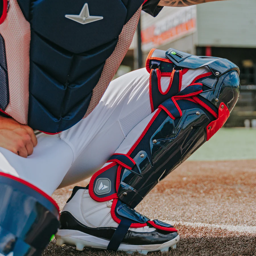 An adult catcher in All Star MVP Pro Catcher's Leg Guards: LG-5 by All Star, featuring a pivoting hinge system in navy and red, crouches behind home plate on the dirt, ready for play.