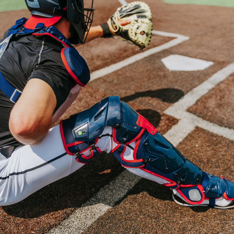 An All Star catcher gears up behind home plate, donning the All Star MVP Pro Catcher's Leg Guards: LG-5 with a pivoting hinge system for one knee down protection, plus helmet, chest protector, and mitt, poised to catch a pitch.